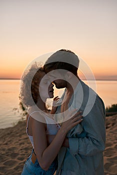 Passionate young couple getting undressed while going to make love, standing on the beach at sunset