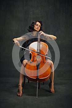 Passionate young woman, musician sitting with eyes closed and playing cello over vintage green background. Being fully