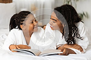 Passtime With Mom. Happy Black Mommy And Daughter Reading Magazine On Bed photo
