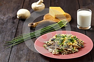 Pasta in a creamy sauce with mushrooms in a plate on a dark table next to a glass of cream, cheese on a wooden board, green onions