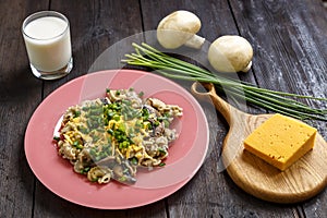 Pasta in a creamy sauce with mushrooms in a plate on a dark wooden table next to a glass of cream, cheese on the board, green