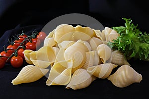 Pasta in the form of shells in a round wooden bowl with cherry tomatoes on a black background. Culinary background