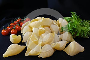 Pasta in the form of shells in a round wooden bowl with cherry tomatoes on a black background. Culinary background