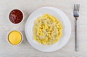 Pasta fusilli in plate, ketchup, mayonnaise, fork on wooden table