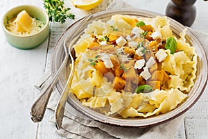 Pasta Mafaldine Napoletane with baked pumpkin, feta cheese and seasoning herbs in ceramic plate on white wooden background.