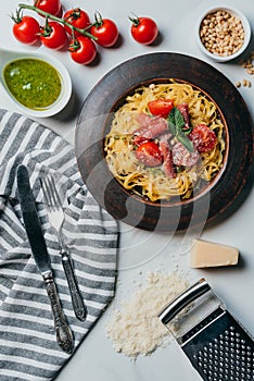 pasta with mint leaves jamon and cherry tomatoes covered by grated parmesan on plate at marble table with kitchen towel knife