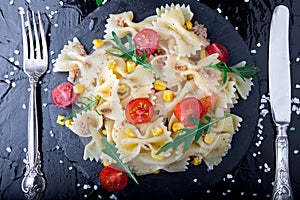 Pasta salad in slate plate with tomatoes cherry, tuna, corn and arugula near knife and spoon. Top view. ingredients. Italian food.