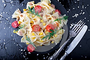 Pasta salad in slate plate with tomatoes cherry, tuna, corn and arugula near knife and spoon. Top view. ingredients. Italian food.