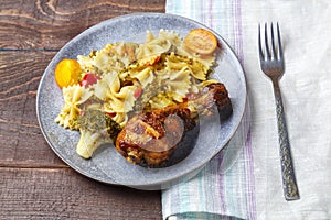 Pasta with vegetables and cherry tomatoes with baked chicken leg on the table next to a light napkin and fork.