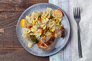 Pasta with vegetables and cherry tomatoes next to a baked chicken leg on the table next to a light napkin and a fork.