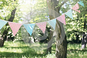 Pastel bunting flags hanging among trees. Summer garden party. Outdoor birthday, wedding decoration. Midsummer, festa