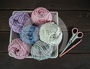 Pastel colored woolen balls in basket with scissors and crochet hook on dark brown wooden ground