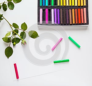 Pastel, pencils, paper and rose flowers placed on white table desk, top view, flat lay.