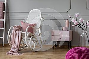 Pastel pink blanket and pillow on white rocking chair in sophisticated room with nightstand and flowers in glass vase, copy space