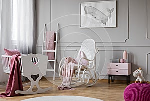 Pastel pink blanket on white rocking chair in spacious baby room interior with cradle, scandinavian ladder and nightstand