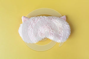 Pastel pink fluffy fur sleep mask with small ears on pastel yellow paper background. Top view, flat lay. Concept of vivid dreams.