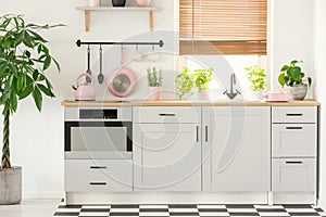 Pastel pink pan and kettle in a beautiful kitchen interior with simple, white cupboards and sink by the window with blinds