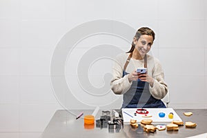 Pastry chef taking picture with phone of cookies while looking at camera