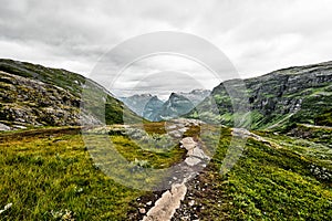 Path over green pasture in the mountains of Western Norway with snow on the summits and a dark cloudy sky