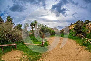 The path among the rocks and green hills leading to the famous beach in Alvor, Algarve, Portugal. Beautiful sky, a bench and a