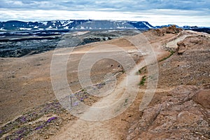 Path to Leirhnjukur old black lava field with colorful stones and smoke coming from ground and blue sky in Iceland, overcast day