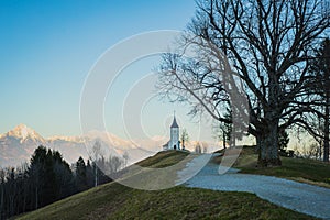 Path towards cute  fantastic charming Saint Primoz church on a small hill with mountains in background at sunset, Jamnik village,