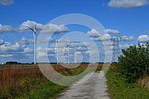 Pathway, high voltage tower with electricity transmission power lines and windmills in field on sunny day