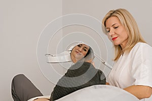 Patient of aesthetic medicine center looks in mirror during consultation with beautician before procedure