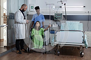 Patient with illness sitting in wheelchair holding drip stand in modern hospital ward