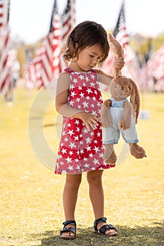 Patriotic holiday child with bunny toy in the park and American flags at Memorial Day