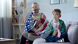 Patriotic old man holding American flag, listening national anthem with grandson