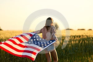 Patriotic woman walking at sunset field, holding with open arms American national flag.