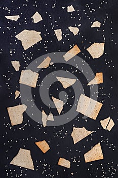 Pattern of pieces of wheat crispbreads with sesame seeds on dark grey shale background.