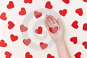 Pattern of red paper hearts surrounding woman`s hand holding two parts of broken heart on the biege background