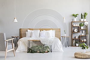 Patterned armchair near white wooden bed in grey bedroom interior with pouf and plants. Real photo