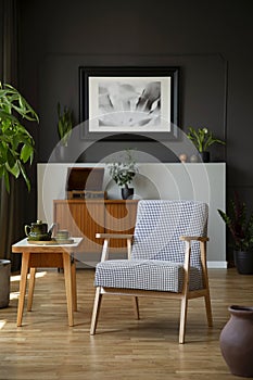 Patterned armchair next to wooden table in dark grey living room interior with poster. Real photo