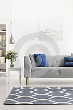 Patterned carpet in front of grey couch with blue pillows in white loft interior with flowers. Real photo