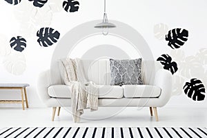 Patterned pillow and blanket on sofa in white living room interior with monstera leaves. Real photo photo