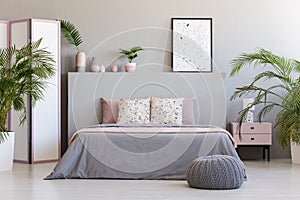 Patterned poster on grey headboard of bed with pillows in bedroom interior with pouf. Real photo