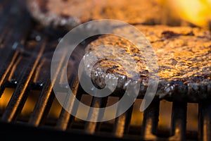 Patties of ground meat with a flame in the background on a cooking grate