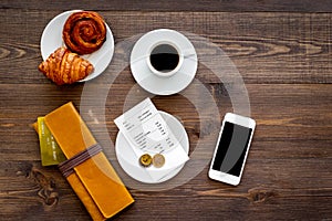 Pay bill at cafe by card. Purse, bill and bank card near coffee and croissant on dark wooden table top view copyspace