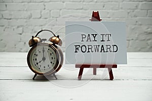 Pay it Forward text and alarm clock on white brick wall and wooden background