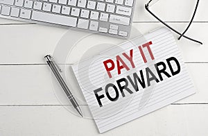 PAY IT FORWARD text on notebook with keyboard , pen glasses on white wooden background