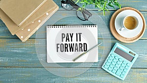 PAY IT FORWARD text on sticky on notebook with pen and glasses , blue background