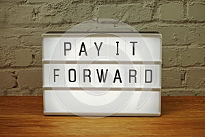 Pay It Forward word in light box on white brick wall and wooden background