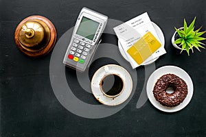 Payment terminal on restaurant desk near bill, service bell, coffee on black background top view copy space