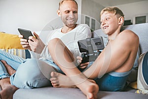 PC games fans Father and son play together with electronic devices, sit in living room on the cozy sofa and smiling cheerfully to