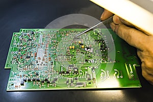 PCB printed circuit board with electrical components and hand with tweezers mounting more parts lit by lamp. Assembling