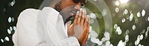 Peaceful African Man Praying In White Shirt Holding Clasped Hands In Prayer Gesture Standing With Eyes Closed Posing In Green