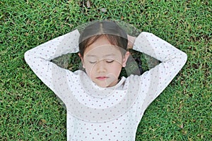 Peaceful Asian little child girl with hands behind head and closed eyes resting on green grass outdoors in summer park. Top view